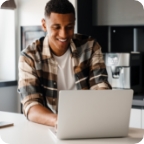 A man stands at a kitchen counter and uses a laptop to compare insurance policies online. 